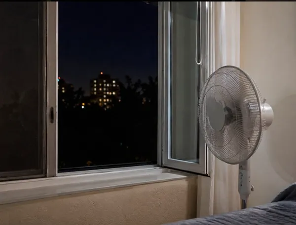 Ventilador encendido junto a ventana abierta de noche para enfriar habitación sin aire acondicionado