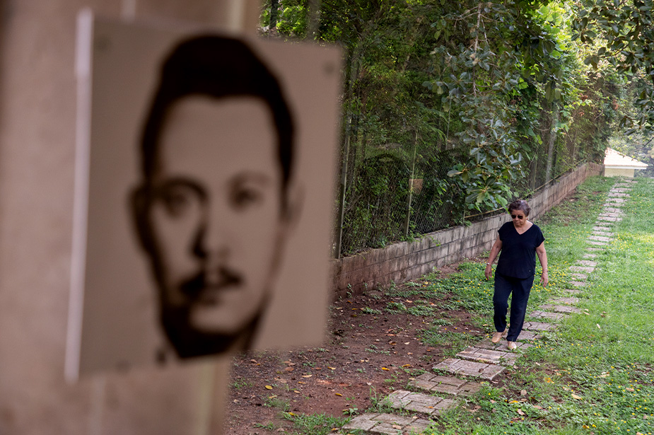 La fotografía de Tomás Nativí, esposo desaparecido de Berta Oliva, enmarca a su fundadora mientras camina hacia él por los jardines del Hogar contra el Olvido, en Santa Ana, Honduras. Nativí fue detenido arbitrariamente y desaparecido el 11 de junio de 1981.